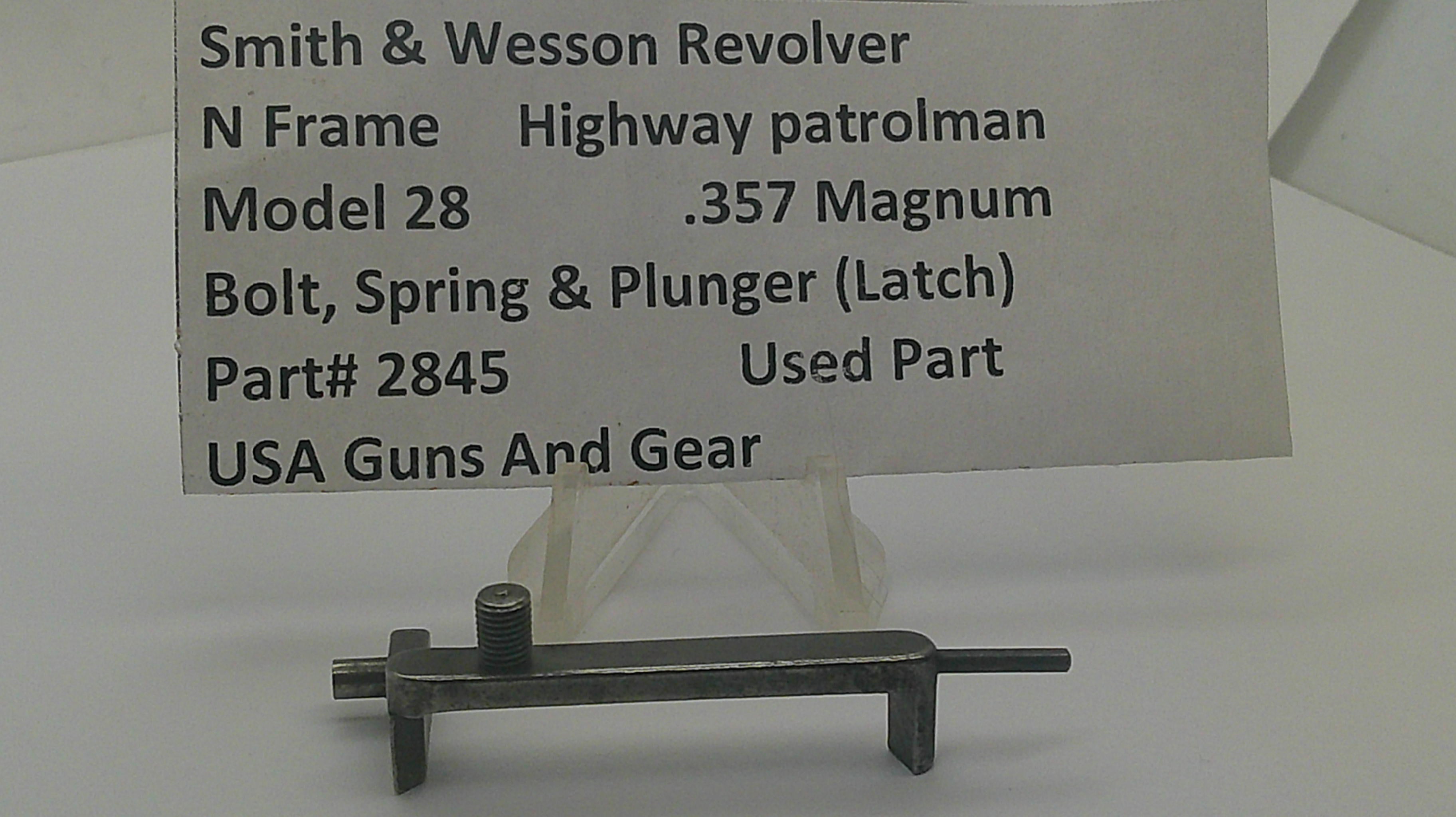 A used metal latch part for a Smith & Wesson revolver, displayed on a clear stand with a paper label card in the background. The label details the part number 2845 and notes it is a used part for a Model 28 .