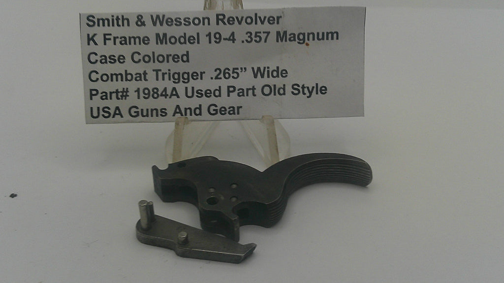 A used black metal Smith & Wesson revolver combat trigger part, displayed on a clear plastic stand in front of a white text label card.