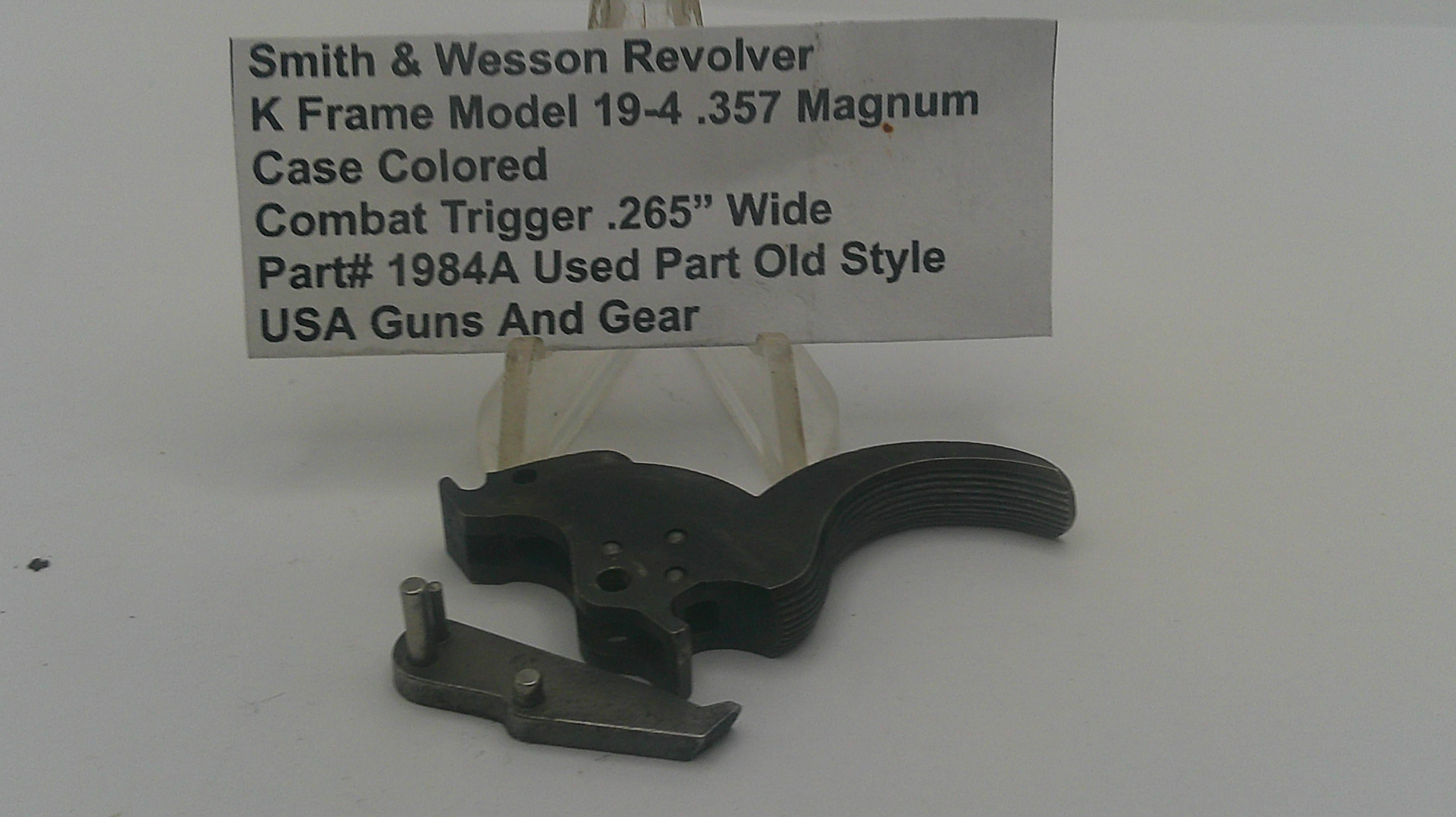 A used black metal Smith & Wesson revolver combat trigger part, displayed on a clear plastic stand in front of a white text label card.