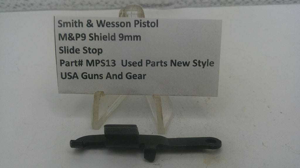 A small black metal slide stop part displayed on a clear plastic stand in front of a white label card that reads 'Smith & Wesson Pistol M&P9 Shield 9mm Slide Stop Part# MPS13 Used Parts New Style USA Guns And Gear'.