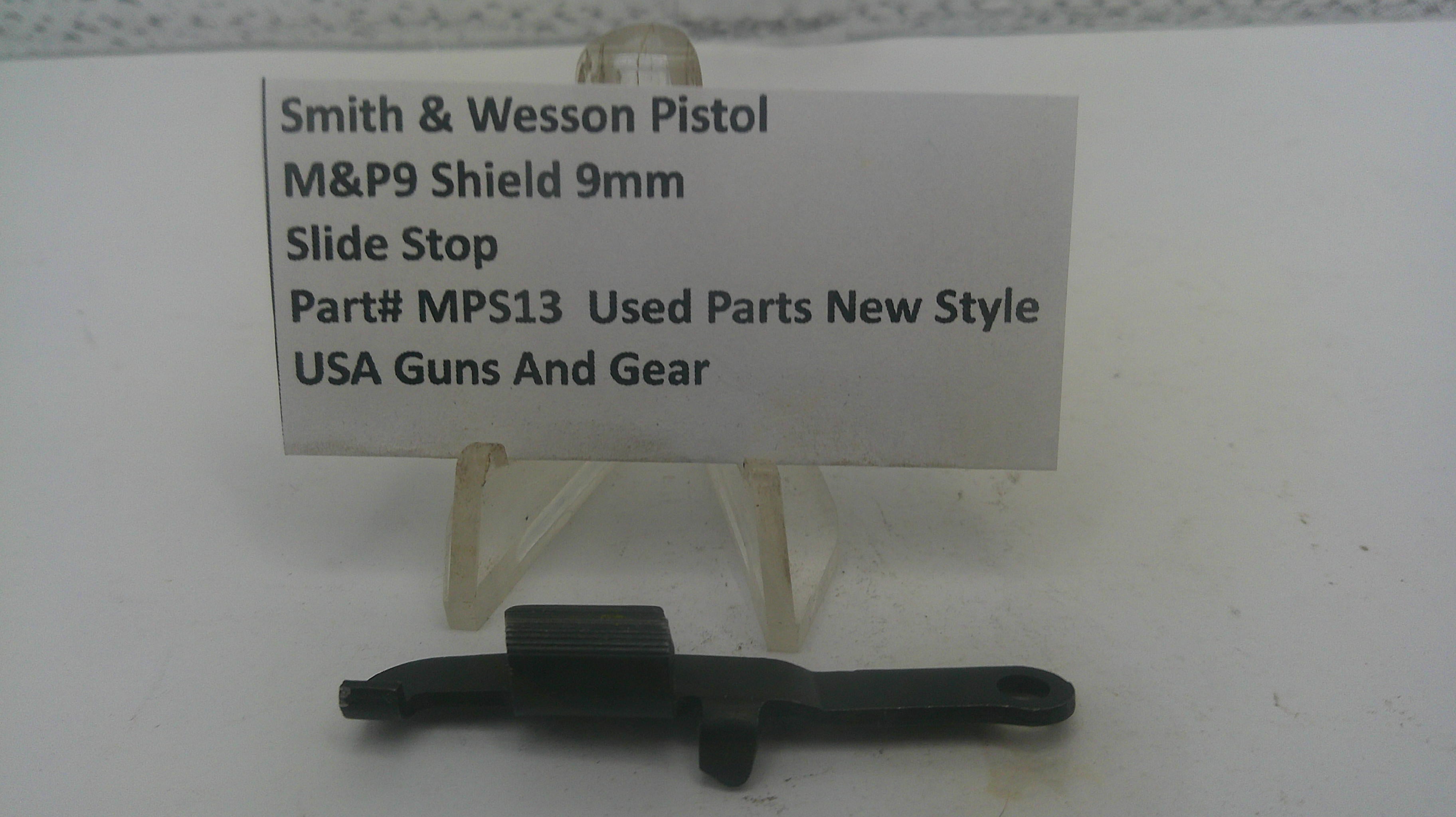 A small black metal slide stop part displayed on a clear plastic stand in front of a white label card that reads 'Smith & Wesson Pistol M&P9 Shield 9mm Slide Stop Part# MPS13 Used Parts New Style USA Guns And Gear'.
