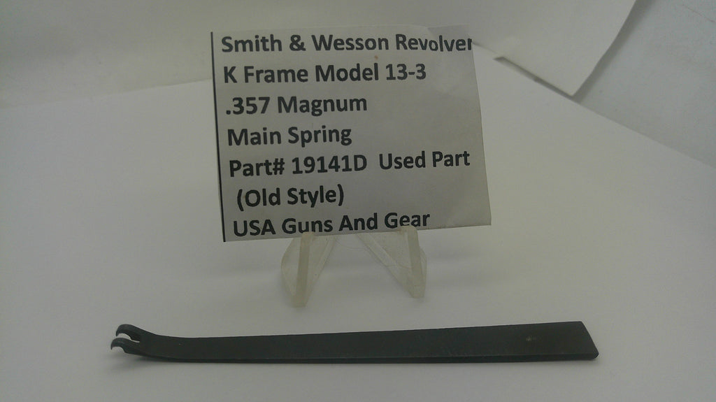 A used black metal main spring part for a Smith & Wesson K-frame revolver, displayed on a white surface with a clear plastic stand holding an identification label.