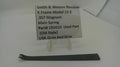 A used black metal main spring part for a Smith & Wesson K-frame revolver, displayed on a white surface with a clear plastic stand holding an identification label.