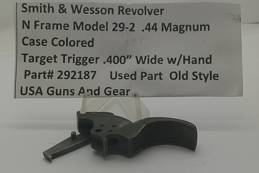 A used Smith & Wesson revolver trigger part, specifically a Model 29-2 .44 Magnum case colored target trigger, displayed on a clear stand in front of a white paper label with product details.