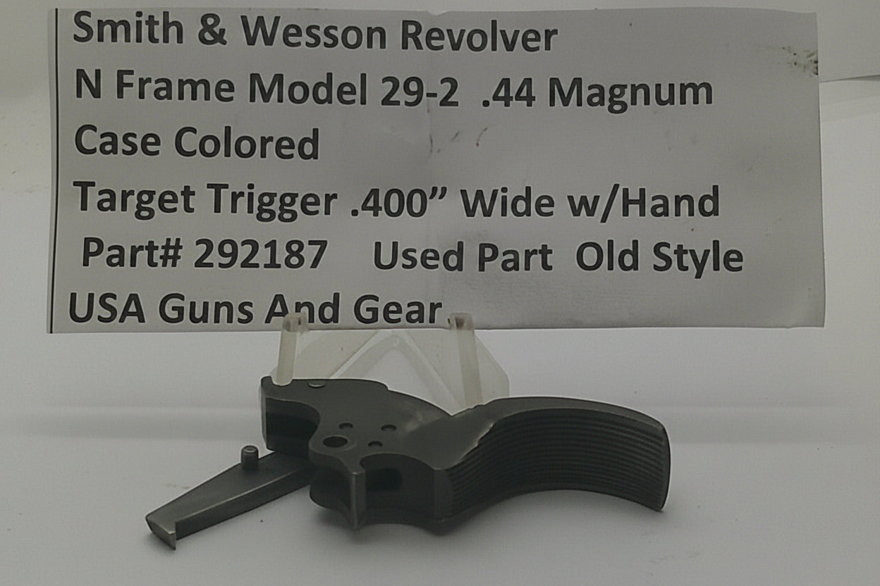 A used Smith & Wesson revolver trigger part, specifically a Model 29-2 .44 Magnum case colored target trigger, displayed on a clear stand in front of a white paper label with product details.