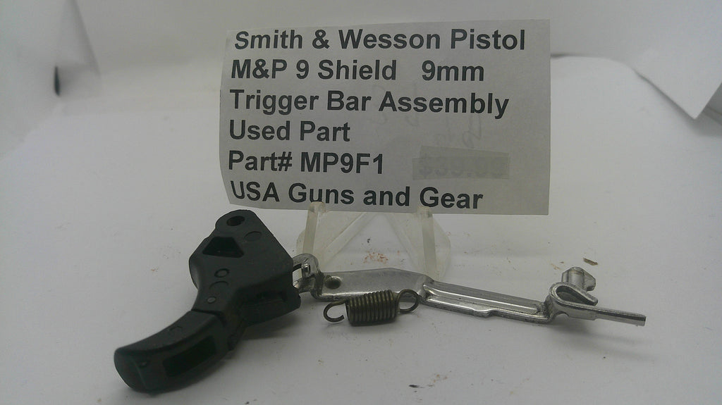 A used Smith & Wesson M&P 9 pistol trigger bar assembly, part number MP9F1, displayed on a white surface with a paper label card behind it.