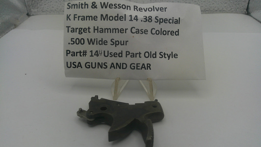 A used black metal Smith & Wesson revolver hammer case part displayed on a small clear stand in front of a white paper label card with product details.