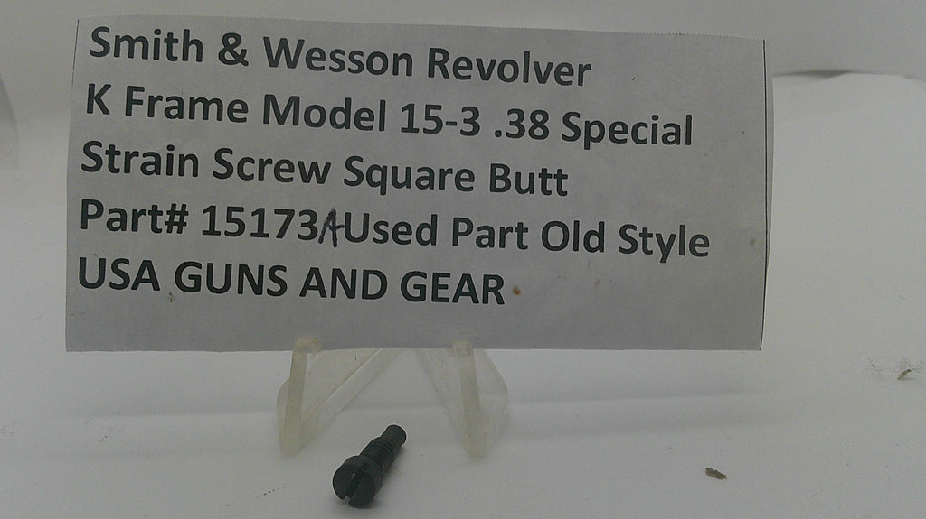 A small black metal strain screw shown on a white surface with a handwritten label card reading 'Smith & Wesson Revolver K Frame Model 15-3 .38 Special Strain Screw Square Butt Part# 15173 Used Part Old Style USA GUNS AND GEAR'.