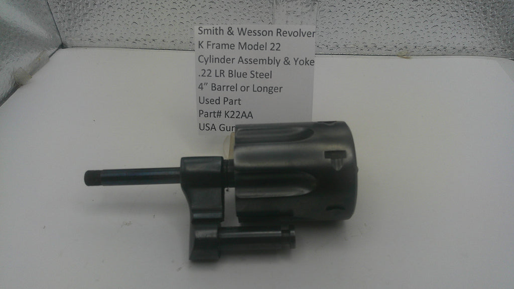 A used black metal Smith & Wesson revolver cylinder assembly and yoke part, shown next to a white paper label with part details, on a white surface.