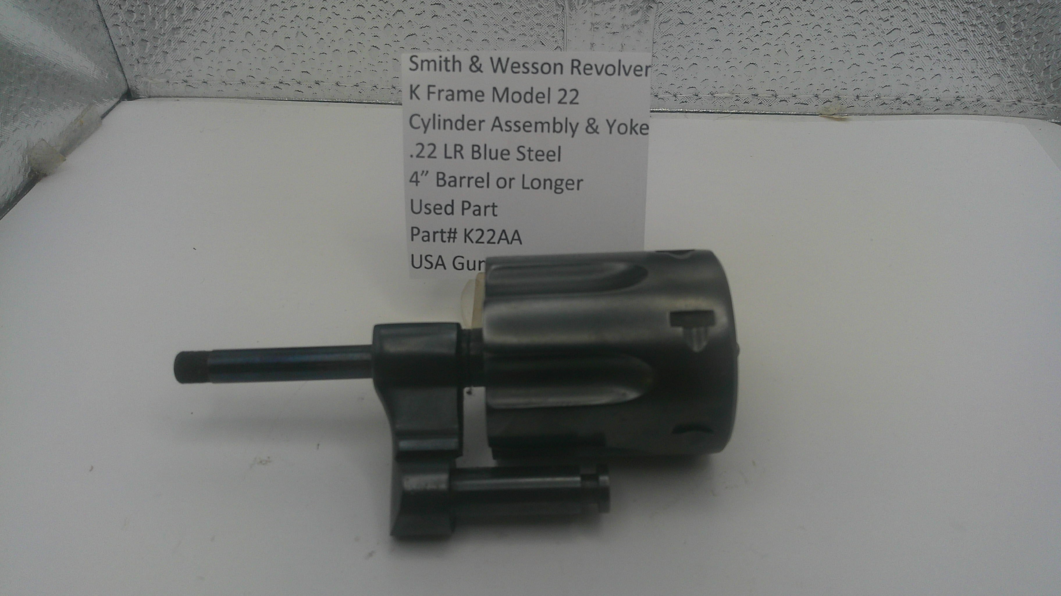 A used black metal Smith & Wesson revolver cylinder assembly and yoke part, shown next to a white paper label with part details, on a white surface.