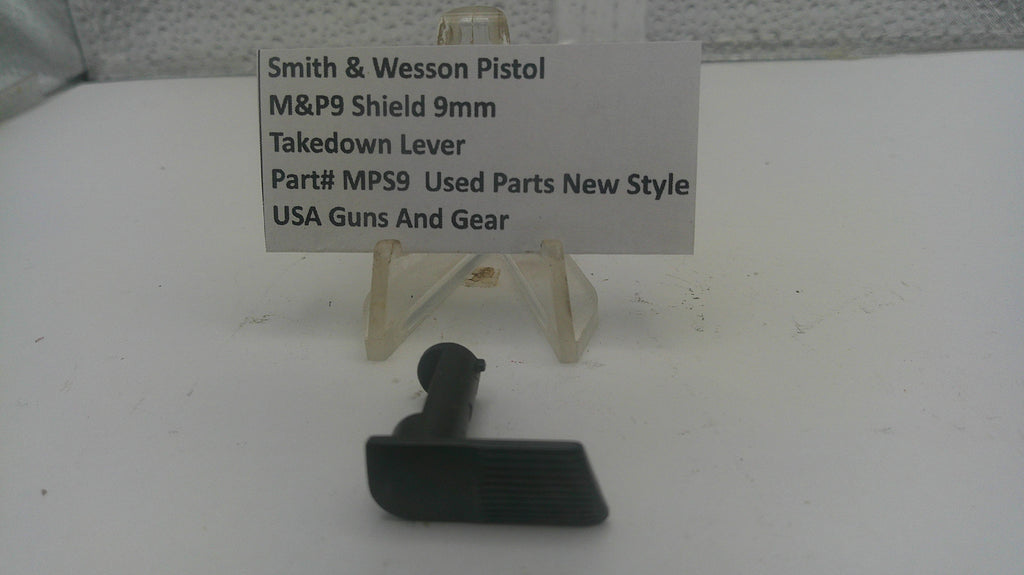 A small black plastic takedown lever part displayed on a white surface in front of a small clear stand with a label card reading 'Smith & Wesson Pistol M&P9 Shield 9mm Takedown Lever Part# MPS9 Used Parts New Style USA Guns And Gear'.