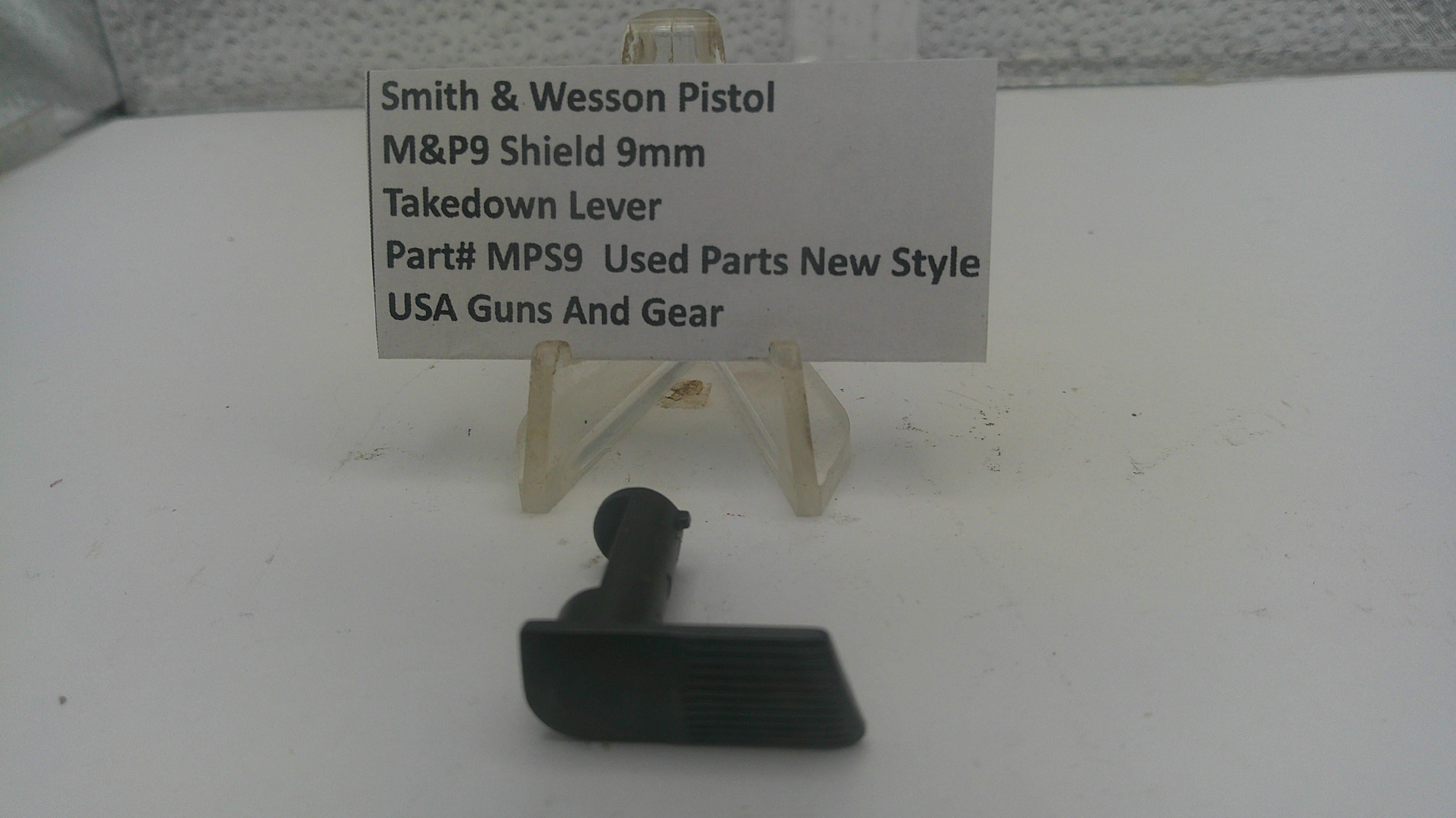 A small black plastic takedown lever part displayed on a white surface in front of a small clear stand with a label card reading 'Smith & Wesson Pistol M&P9 Shield 9mm Takedown Lever Part# MPS9 Used Parts New Style USA Guns And Gear'.