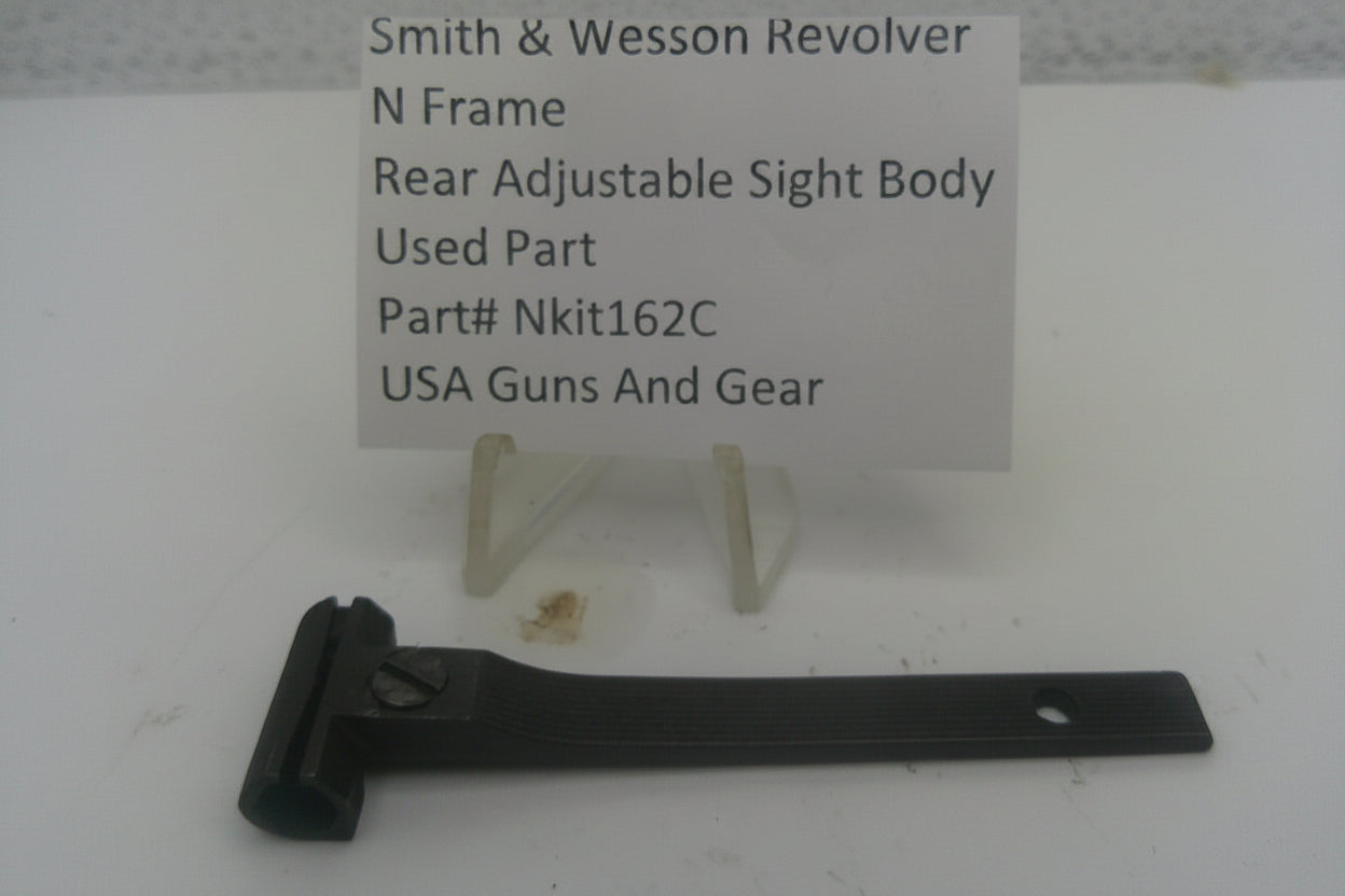 A black metal rear adjustable sight body part for a Smith & Wesson revolver, displayed on a white surface with a small sign behind it reading 'Smith & Wesson Revolver N Frame Rear Adjustable Sight Body Used Part Part# Nkit162A USA Guns And Gear'.