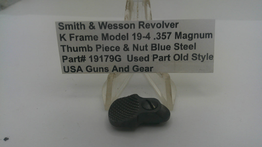 A used black thumb piece and nut assembly for a Smith & Wesson revolver, displayed on a clear plastic stand with a white label card in the background.