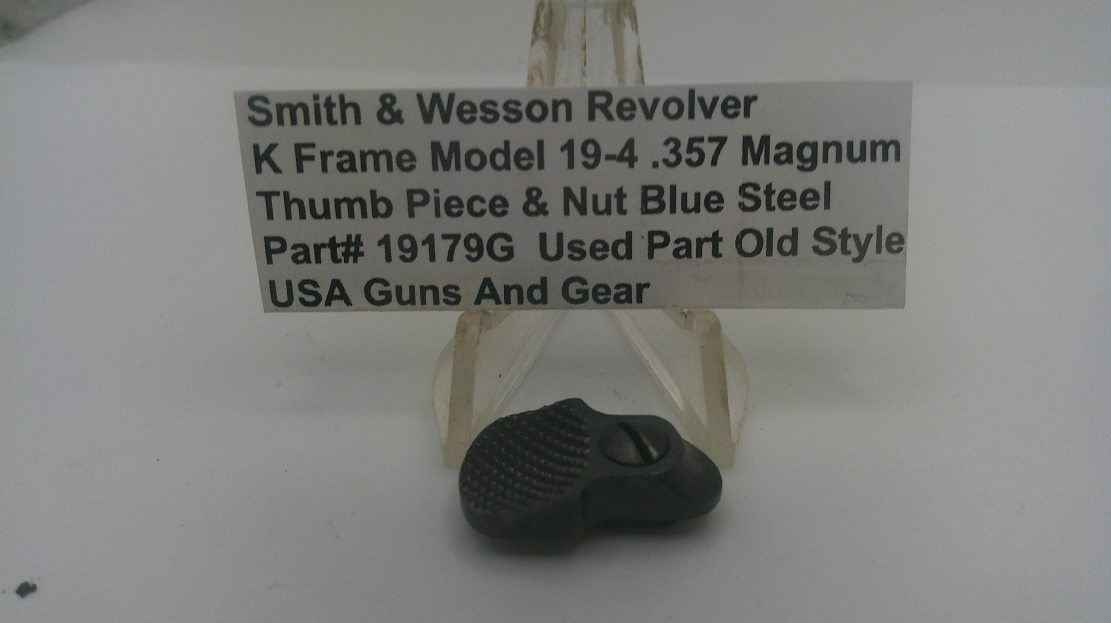 A used black thumb piece and nut assembly for a Smith & Wesson revolver, displayed on a clear plastic stand with a white label card in the background.