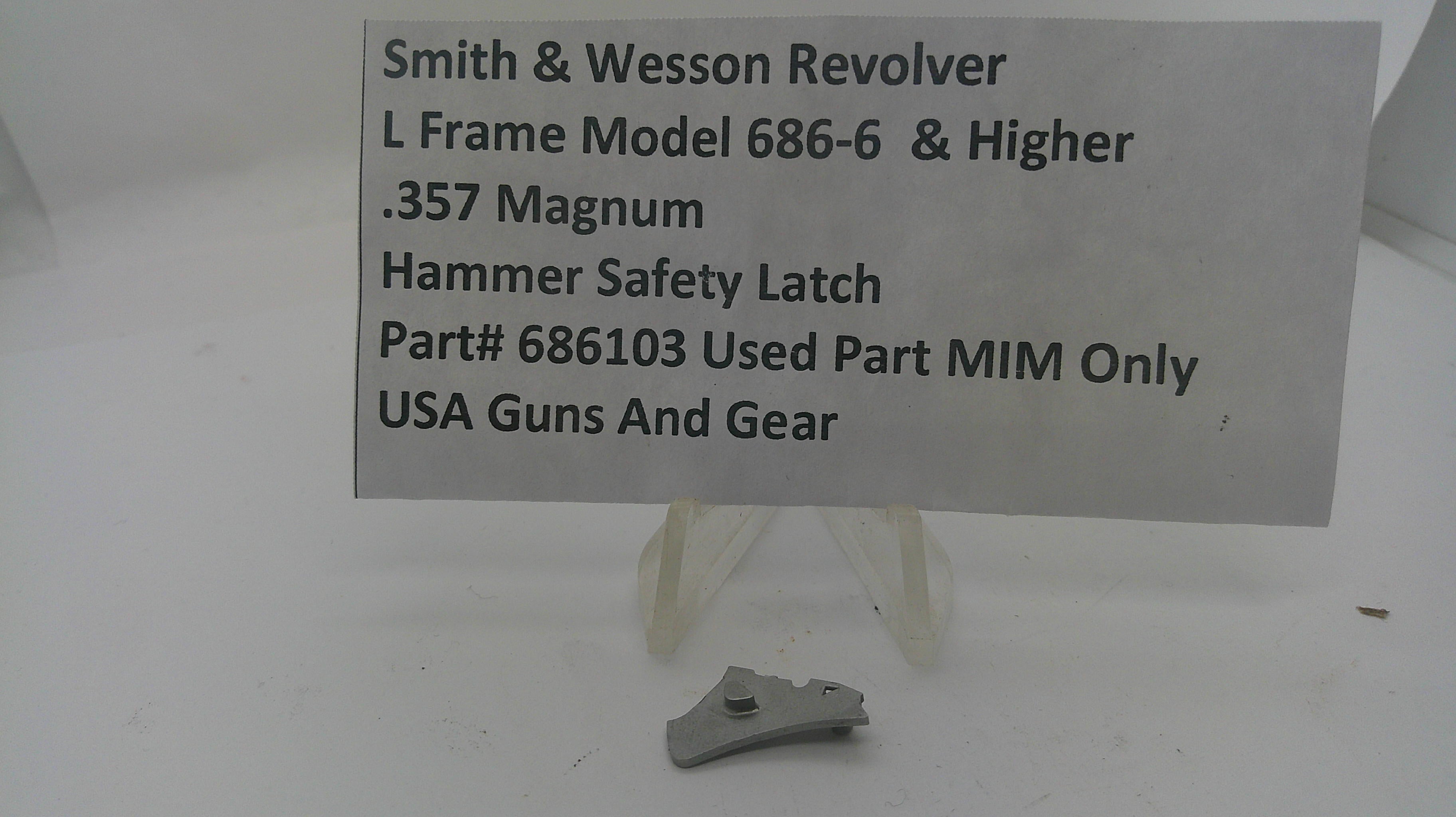 A small metal hammer safety latch part displayed on a clear stand beneath a printed label card that reads 'Smith & Wesson Revolver L Frame Model 686-6 & Higher .357 Magnum Hammer Safety Latch Part# 686103 Used Part MIM Only USA Guns And Gear'.