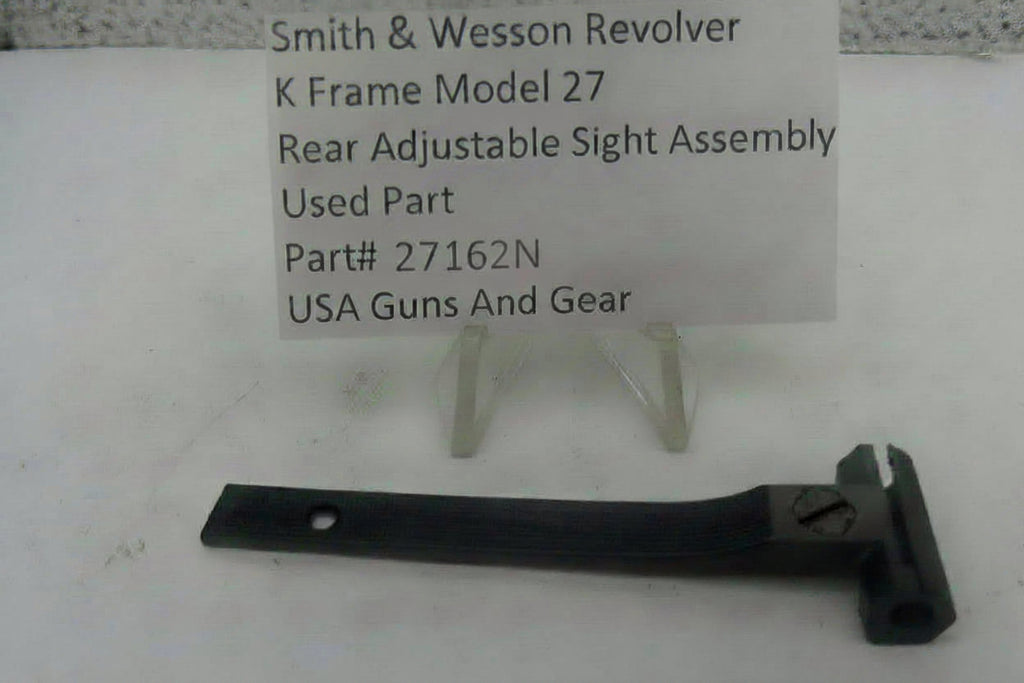 A used black metal rear adjustable sight assembly for a Smith & Wesson revolver, displayed on a white surface with a white paper label card on small plastic stands.