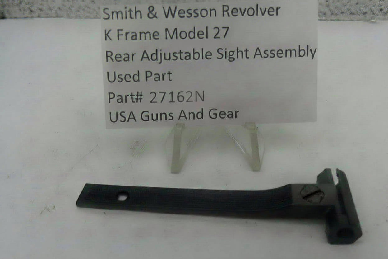 A used black metal rear adjustable sight assembly for a Smith & Wesson revolver, displayed on a white surface with a white paper label card on small plastic stands.