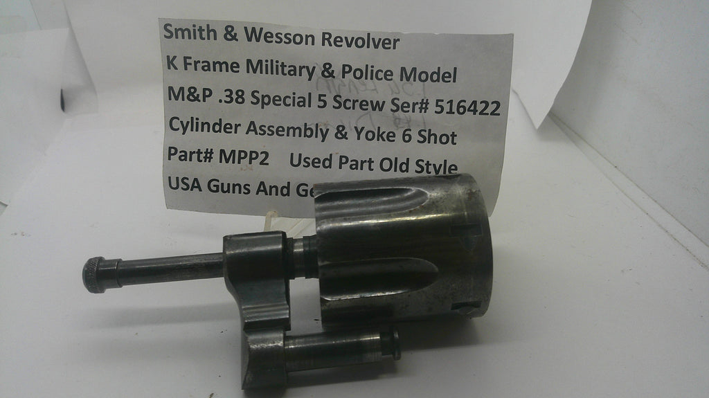 A used metal Smith & Wesson revolver cylinder assembly and yoke 6 shot part, shown with a paper label card in the background on a white surface.