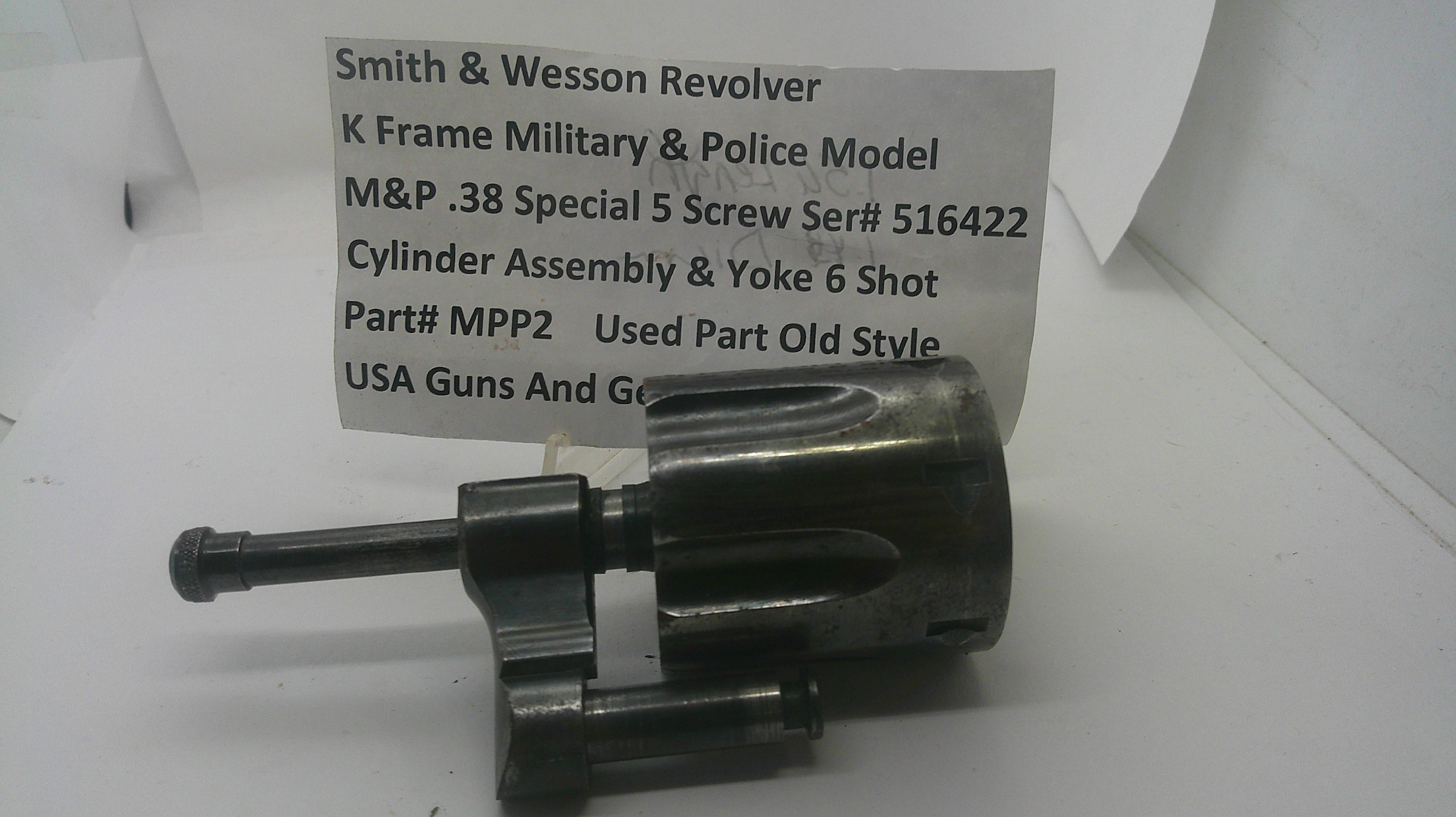 A used metal Smith & Wesson revolver cylinder assembly and yoke 6 shot part, shown with a paper label card in the background on a white surface.