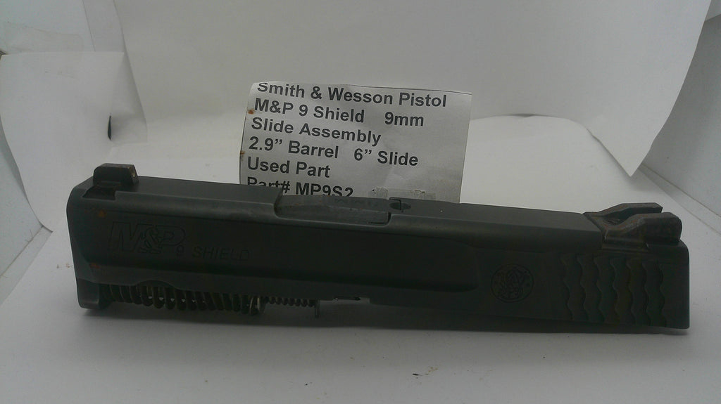 A used black Smith & Wesson pistol slide assembly, part number MP932, displayed on a white surface with a white paper label card behind it.