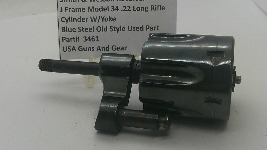 A used black metal revolver cylinder and frame part, photographed on a white surface with a paper label in the background reading Smith & Wesson J Frame Model 34 .22 Long Rifle Cylinder W/Yoke Blue Steel Old Style Used Part Part# 3461 USA Guns And Gear.