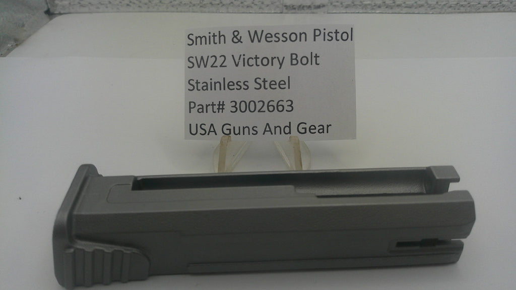A dark gray metal receiver or grip component for a Smith & Wesson pistol, displayed on a white surface with a paper label card behind it reading 'Smith & Wesson Pistol SW22 Victory Bolt Stainless Steel Part# 3002663 USA Guns And Gear'.
