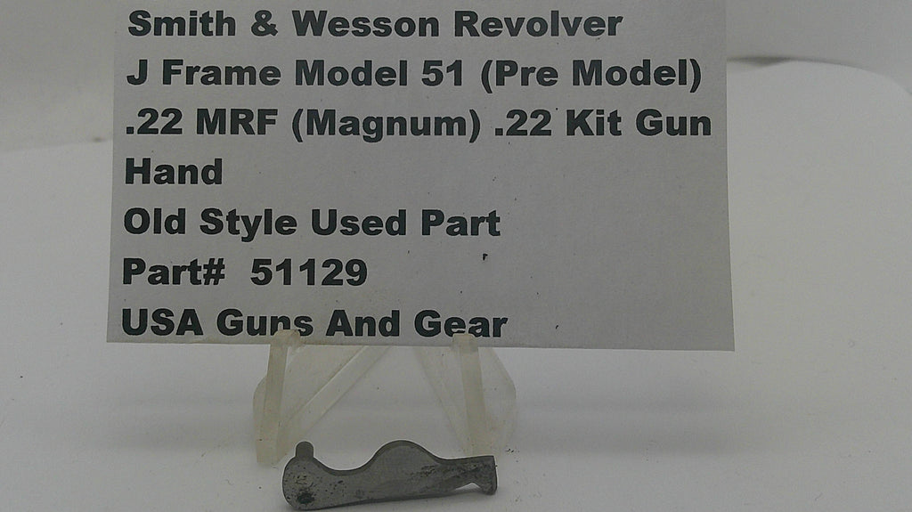 A small metal used part for a Smith & Wesson revolver, displayed on a clear plastic stand in front of a white sign with product details.