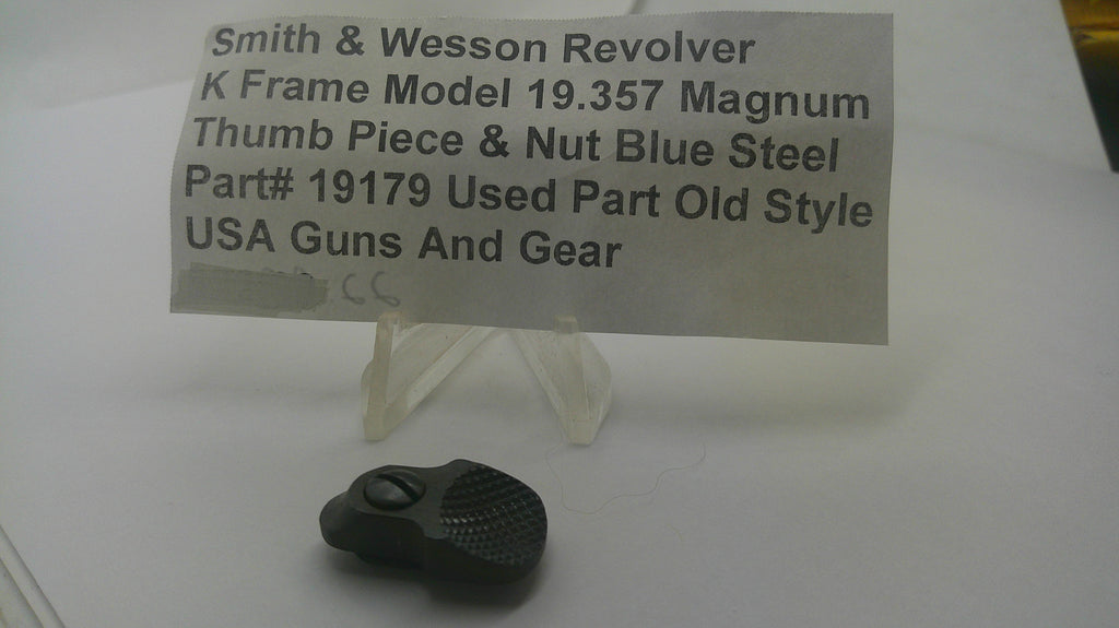 A small black textured metal part displayed on a clear plastic stand in front of a paper label that reads 'Smith & Wesson Revolver K Frame Model 19.357 Magnum Thumb Piece & Nut Blue Steel Part# 19179 Used Part Old Style USA Guns And Gear'.