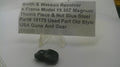 A small black textured metal part displayed on a clear plastic stand in front of a paper label that reads 'Smith & Wesson Revolver K Frame Model 19.357 Magnum Thumb Piece & Nut Blue Steel Part# 19179 Used Part Old Style USA Guns And Gear'.