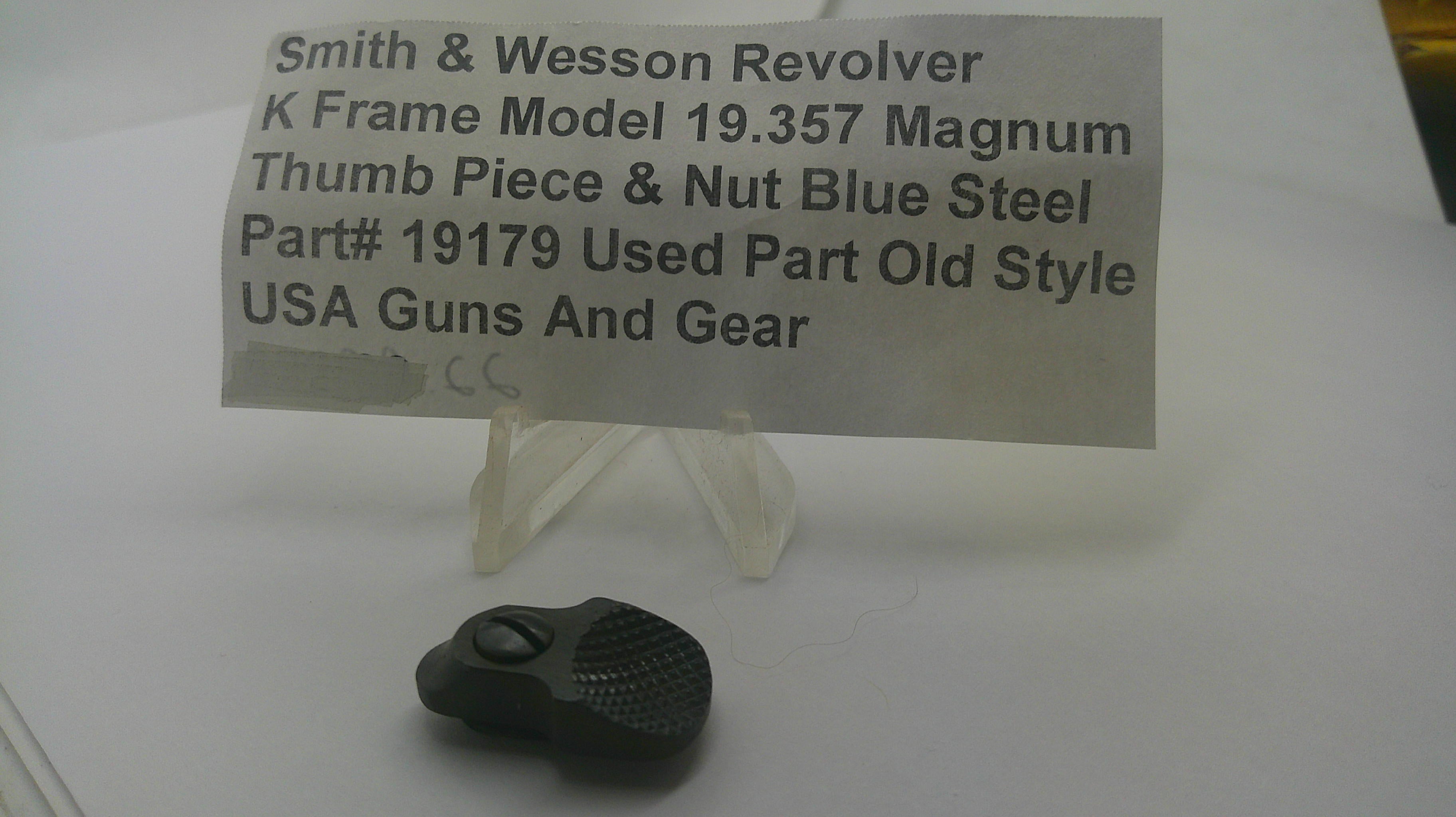 A small black textured metal part displayed on a clear plastic stand in front of a paper label that reads 'Smith & Wesson Revolver K Frame Model 19.357 Magnum Thumb Piece & Nut Blue Steel Part# 19179 Used Part Old Style USA Guns And Gear'.