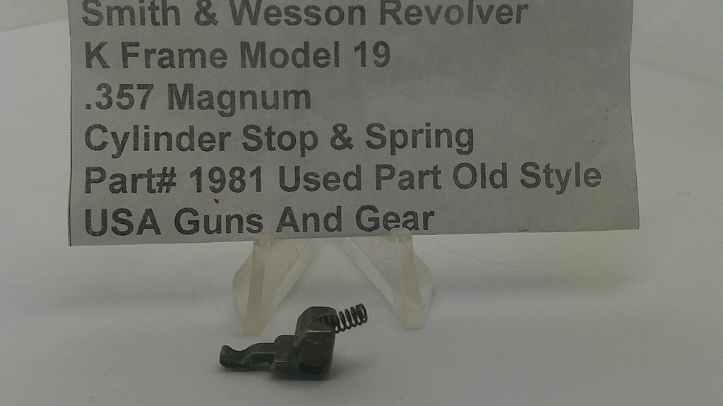A small metal cylinder stop and spring part displayed on a clear plastic stand, with a paper label reading 'Smith & Wesson Revolver K Frame Model 19 .357 Magnum Cylinder Stop & Spring Part# 1981 Used Part Old Style USA Guns And Gear' in the background.