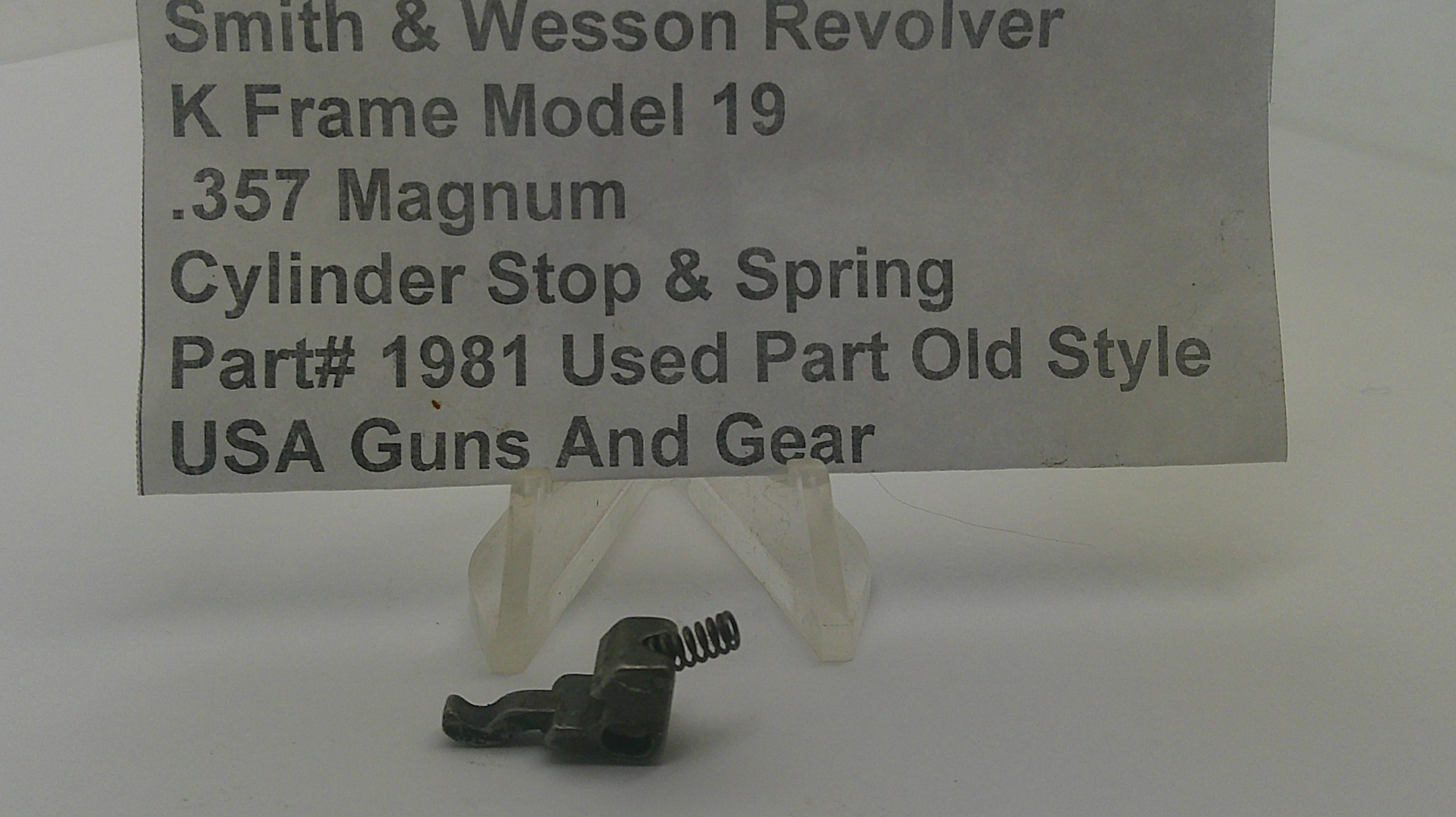 A small metal cylinder stop and spring part displayed on a clear plastic stand, with a paper label reading 'Smith & Wesson Revolver K Frame Model 19 .357 Magnum Cylinder Stop & Spring Part# 1981 Used Part Old Style USA Guns And Gear' in the background.