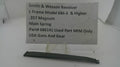 A used black metal main spring for a Smith & Wesson revolver, displayed on a clear plastic stand with a white paper label showing the part number and model details.