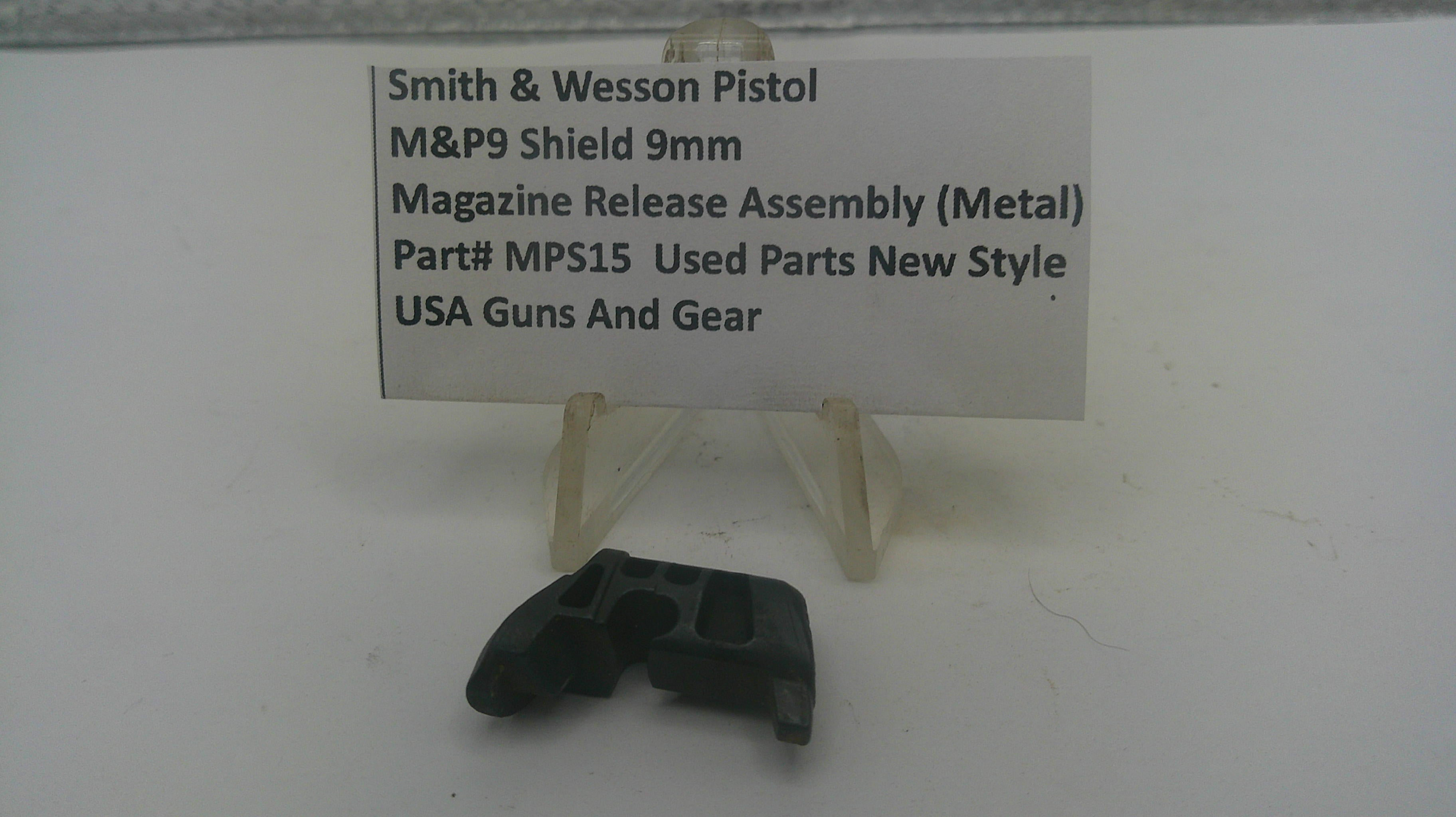 A small black metal magazine release assembly part displayed on a clear plastic stand in front of a white sign that reads 'Smith & Wesson Pistol M&P9 Shield 9mm Magazine Release Assembly (Metal) Part# MPS15 Used Parts New Style USA Guns And Gear'.
