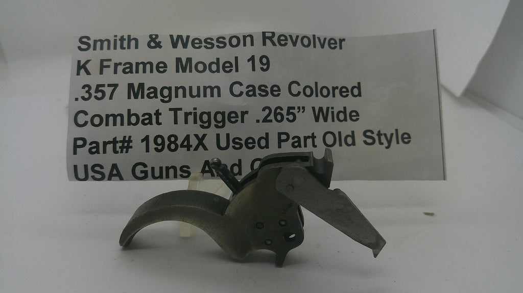 A used metal Smith & Wesson revolver trigger component, model K Frame Model 19, shown in front of a white paper label with product details.