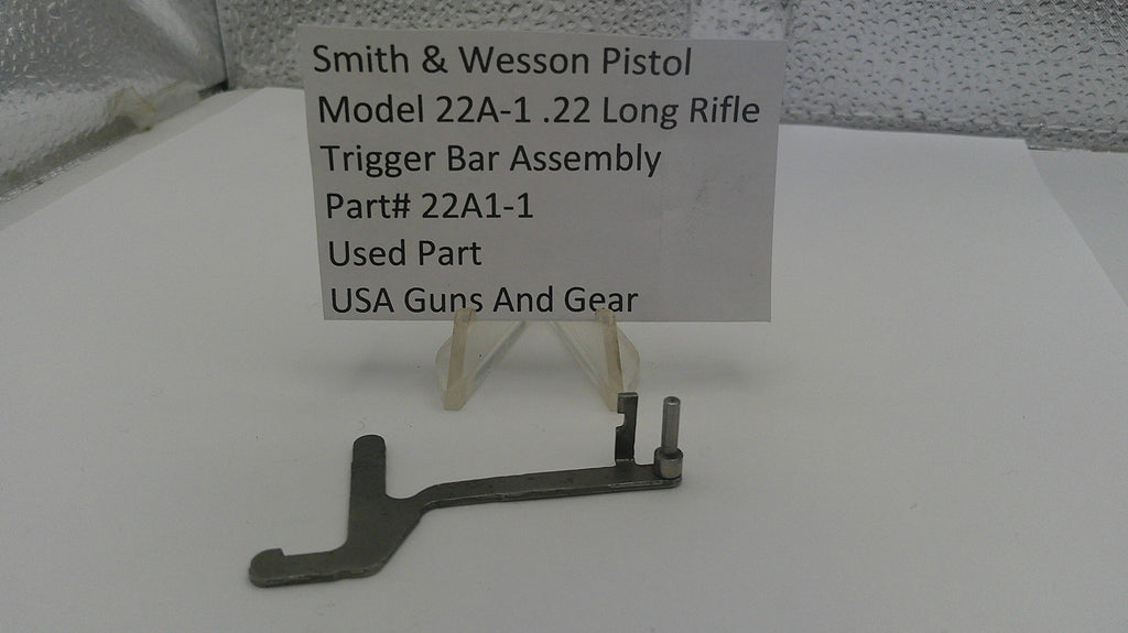 Smith & Wesson pistol trigger bar assembly on a white surface with a label.
