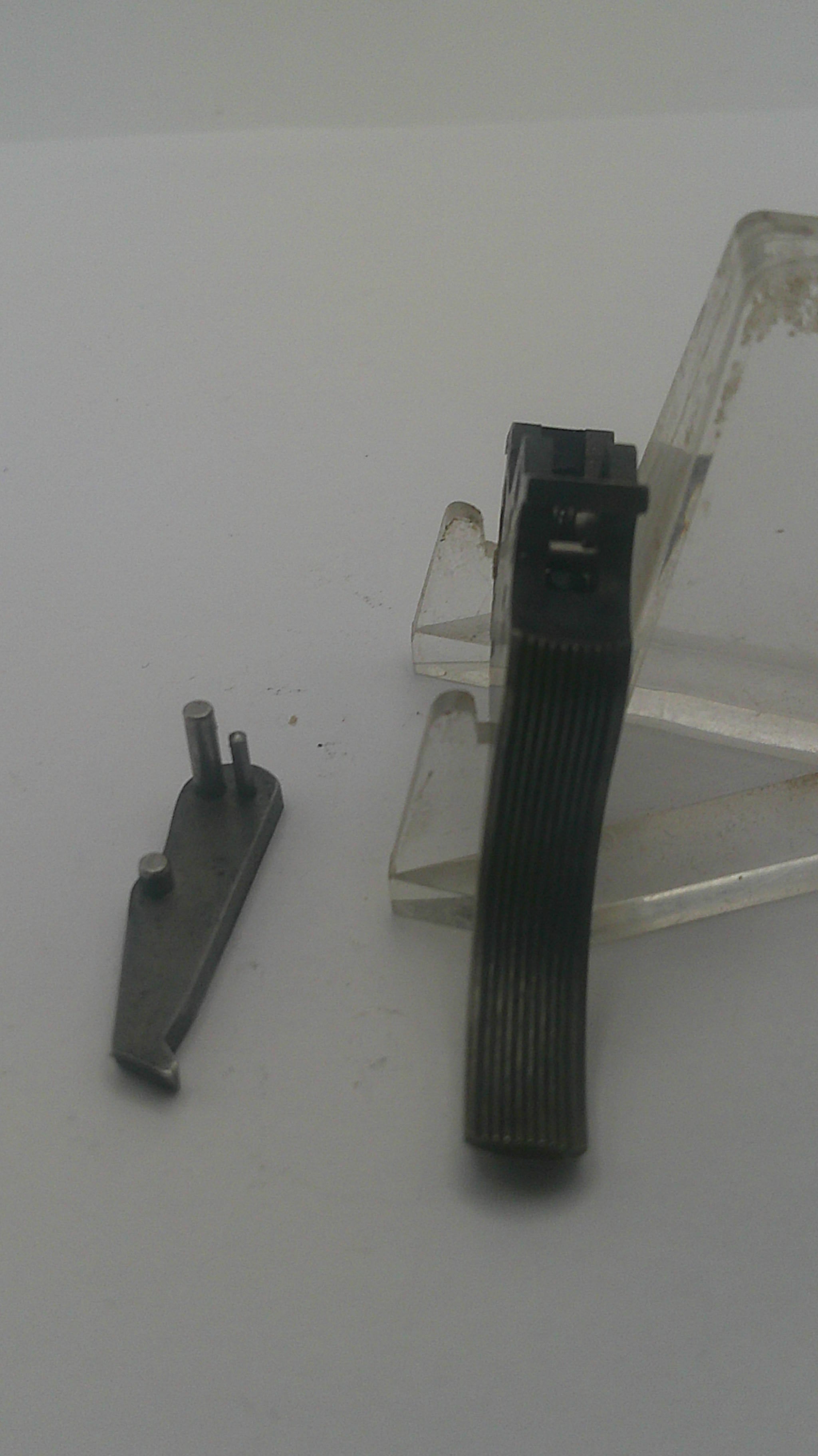 A small dark metal hardware component with a flat ribbed black strip across the middle, shown against a plain light gray background with a clear plastic stand.