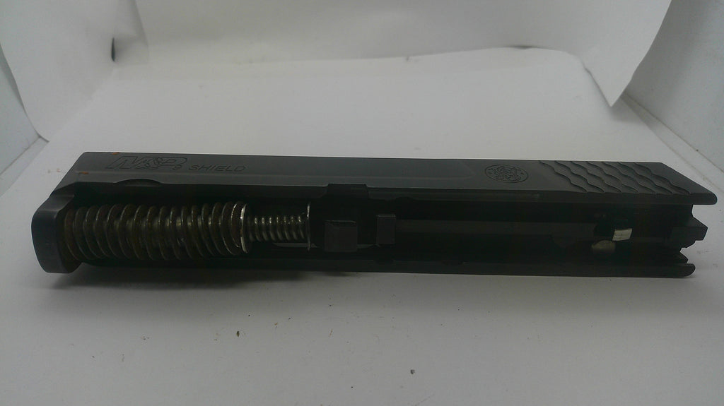 A black metal firearm accessory, likely a receiver or grip component with a visible coiled spring, photographed on a white background.