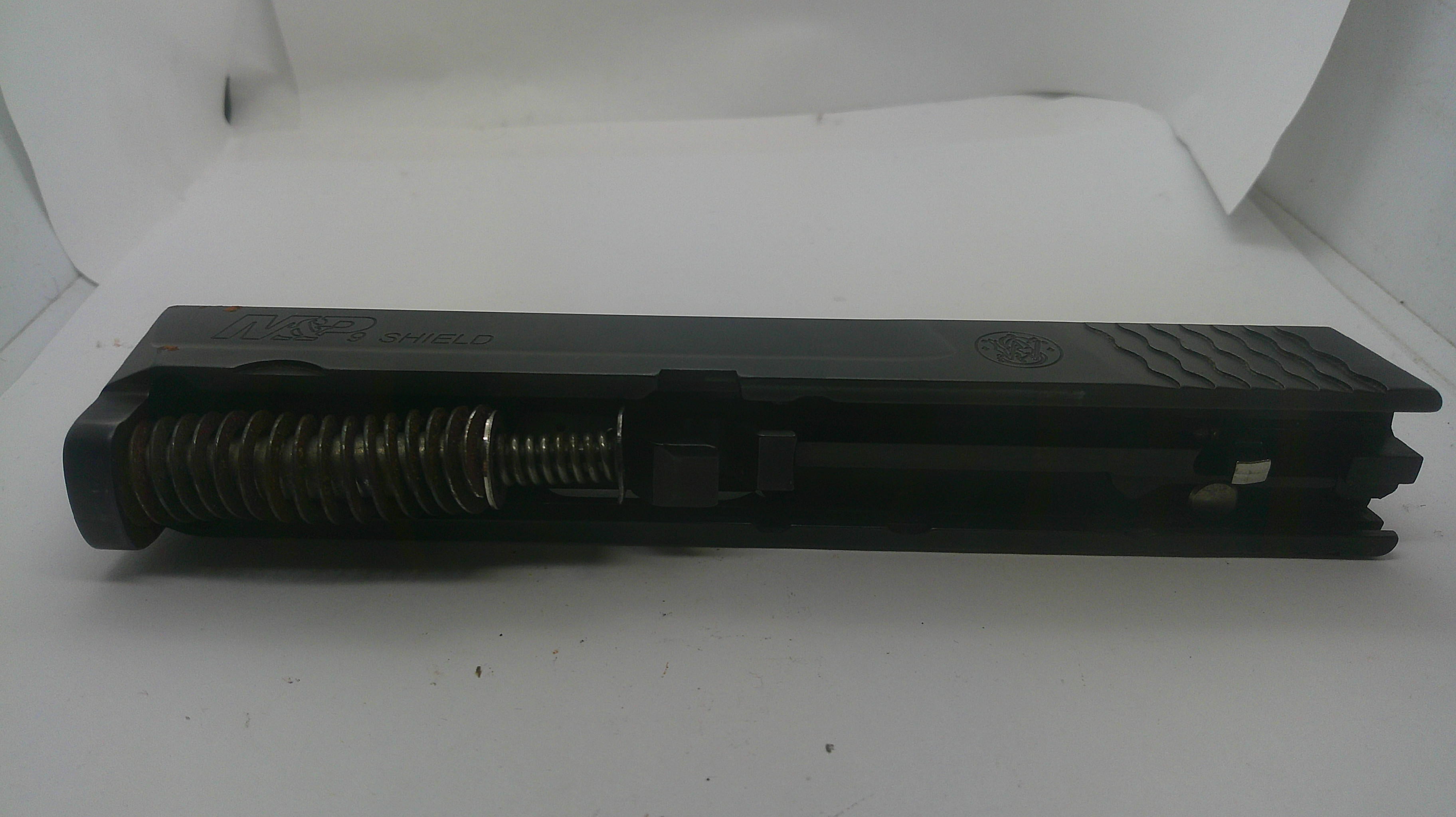A black metal firearm accessory, likely a receiver or grip component with a visible coiled spring, photographed on a white background.