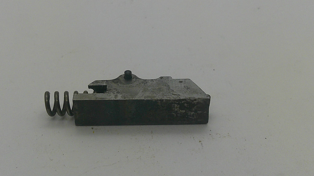 A small dark metal mechanical component with a coiled spring attached on the left side, photographed on a plain light background.