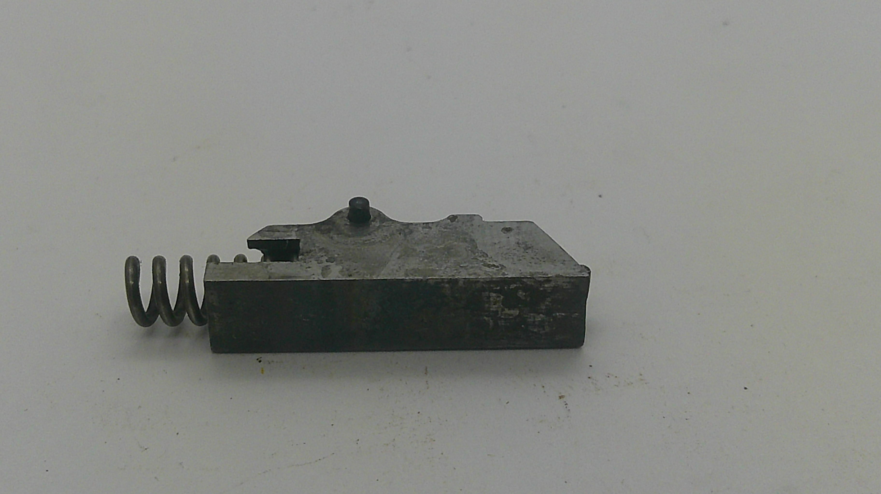 A small dark metal mechanical component with a coiled spring attached on the left side, photographed on a plain light background.