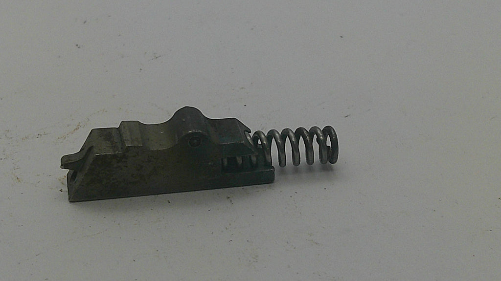 A small dark metal mechanical component with a coiled spring attached, photographed on a plain off-white surface.