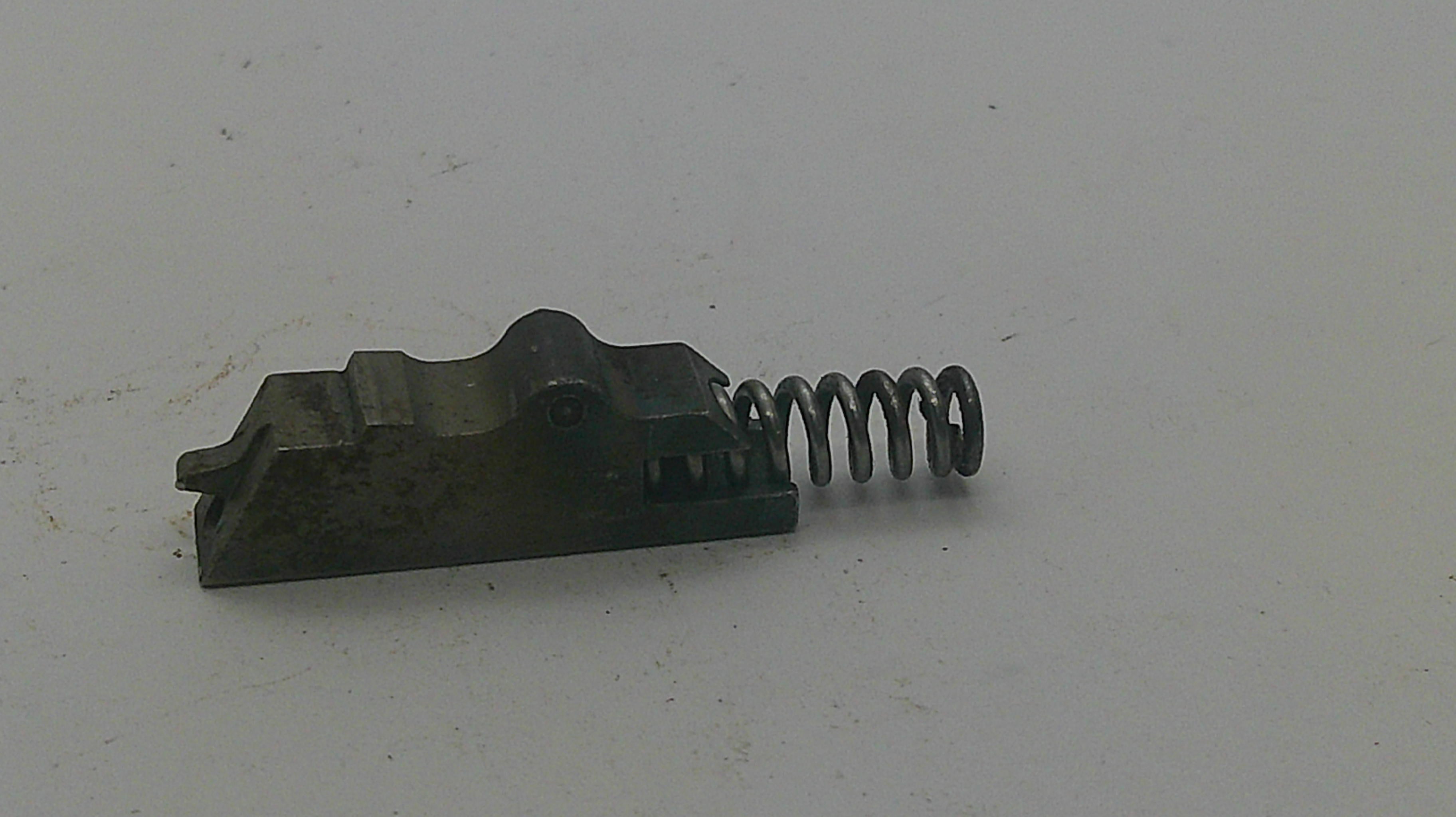 A small dark metal mechanical component with a coiled spring attached, photographed on a plain off-white surface.