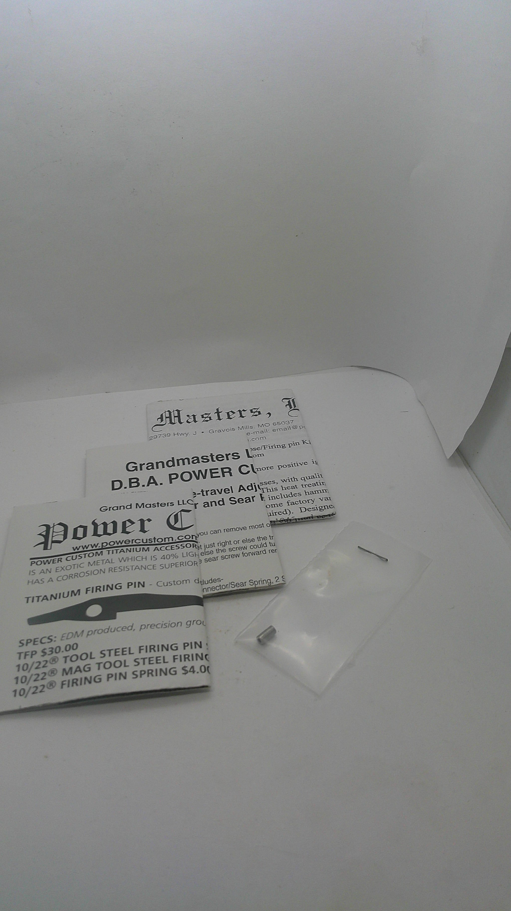 A white cardboard box containing printed inserts and a small clear plastic bag with a metal pin, photographed on a white background.