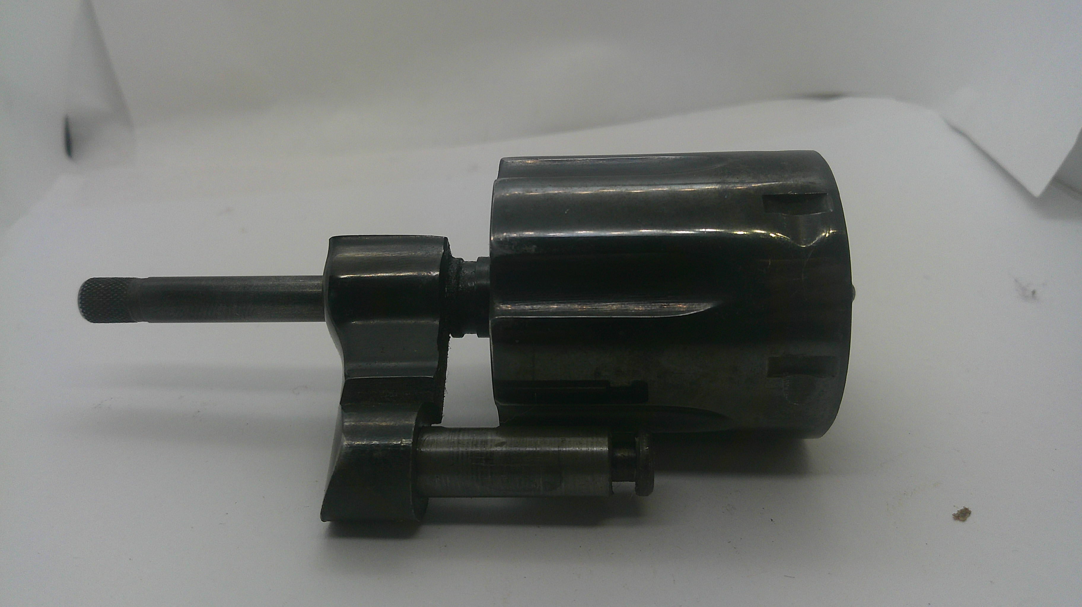 A dark metallic cylindrical mechanical part with a protruding rod and small attached cylindrical component, photographed on a plain white surface.