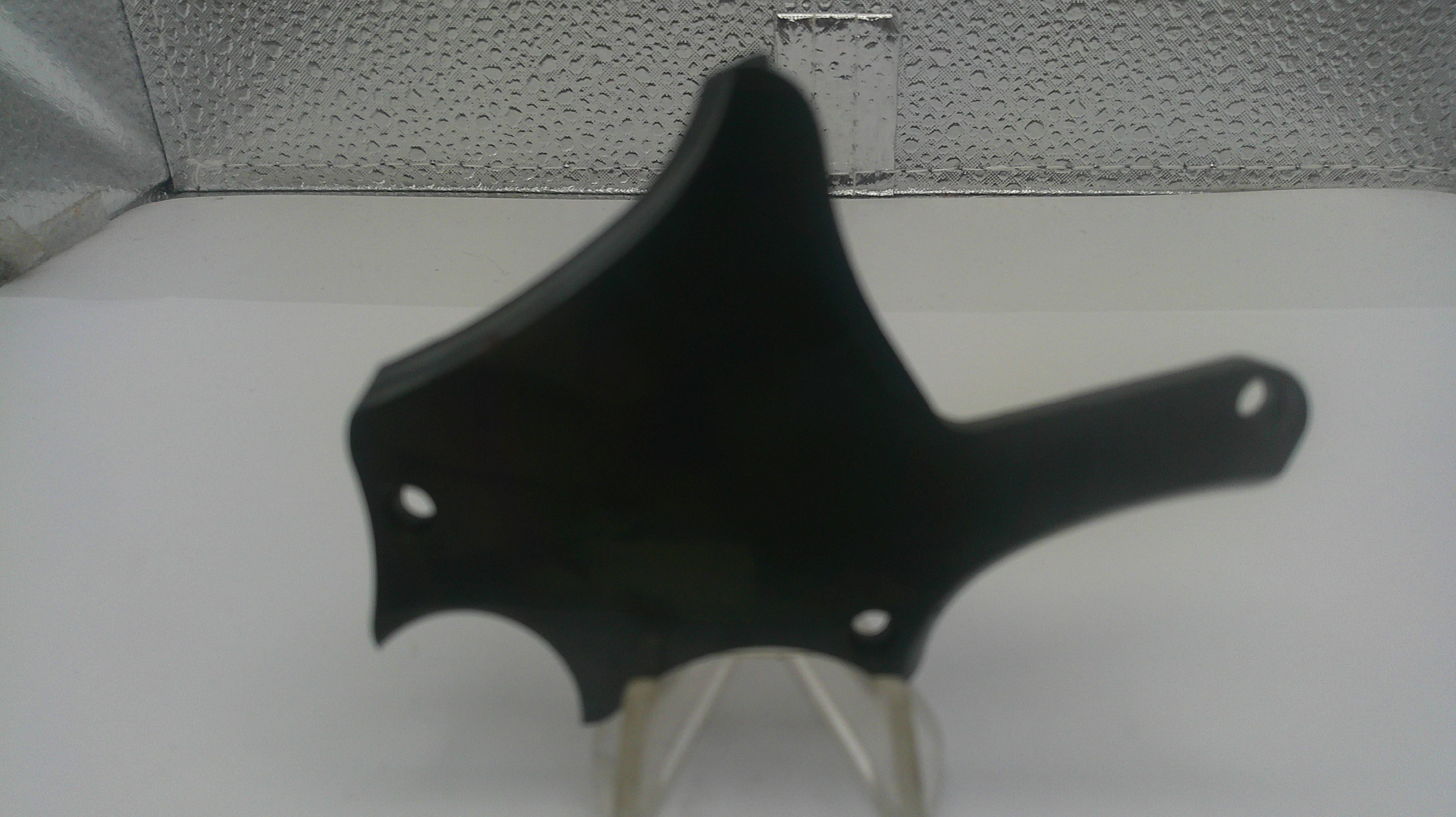A black plastic motorcycle or scooter handlebar trim piece with mounting holes, displayed on a clear stand against a white surface and textured gray background.