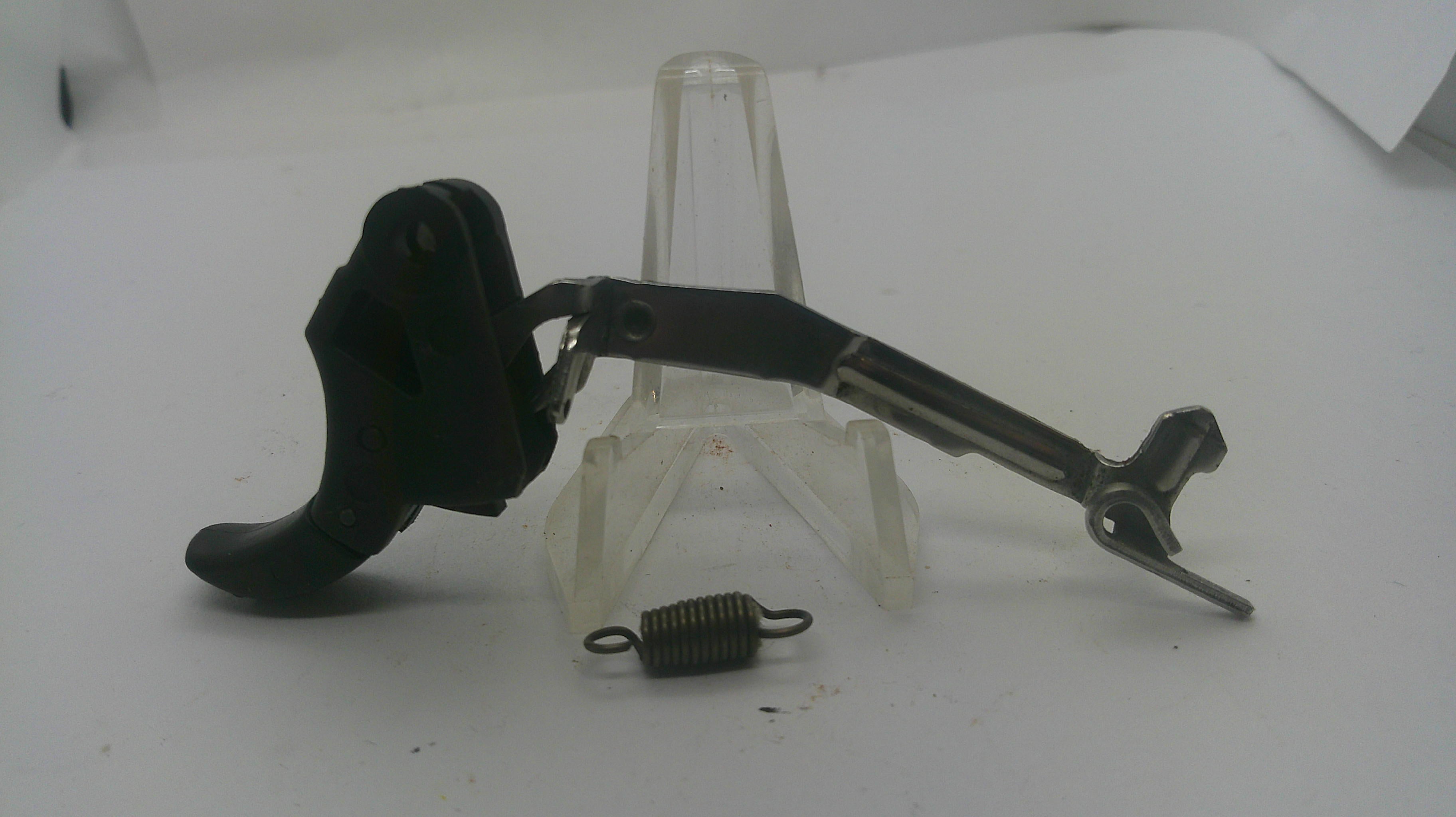 A replacement trigger assembly for a firearm, featuring a black pistol grip, a silver metal lever, and a small coiled spring, displayed on a clear plastic stand against a white background.