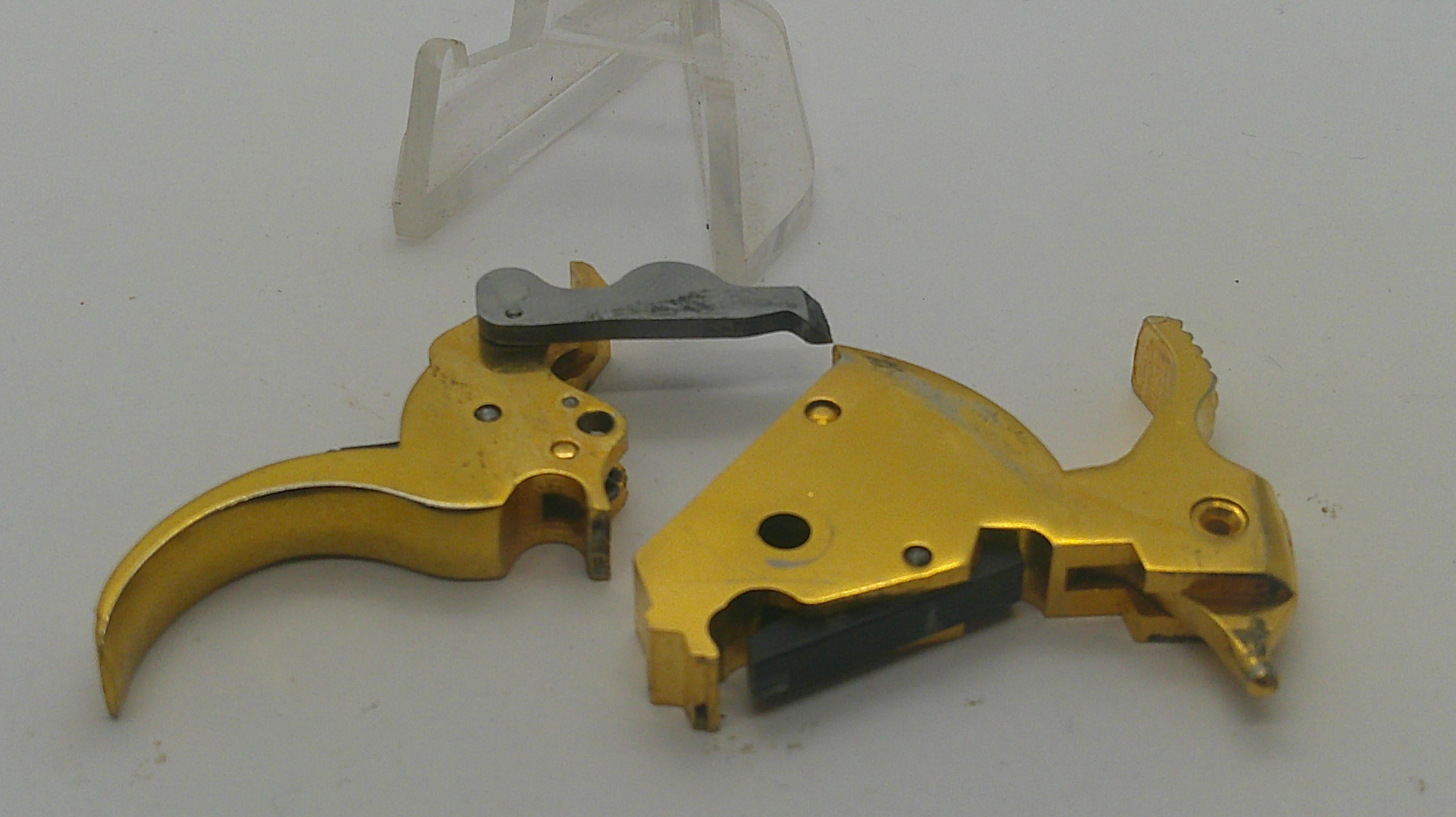 Gold-finished pistol trigger assembly components, including a gold receiver and a silver-colored hammer, shown on a light gray background with a clear plastic stand in the background.