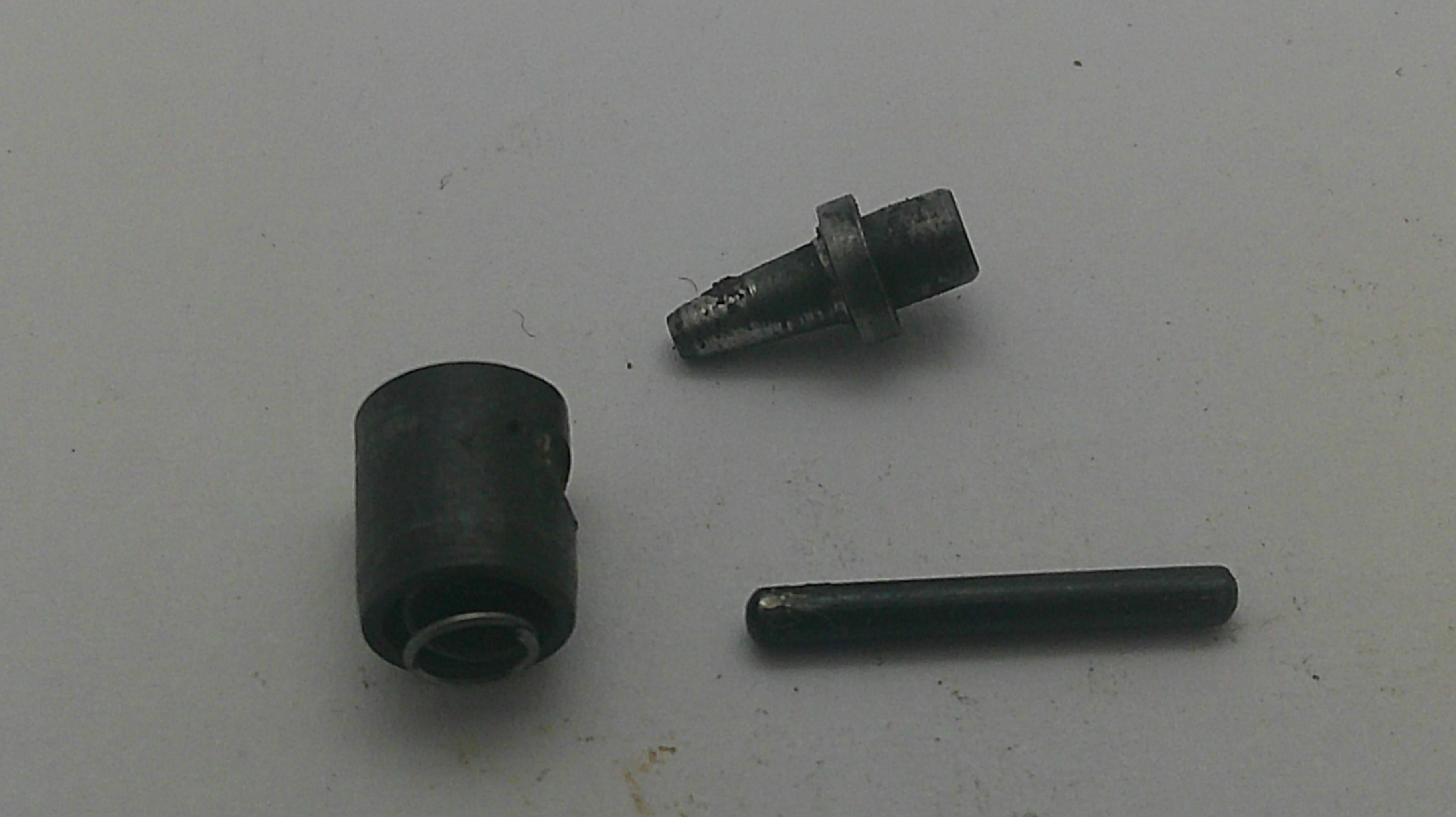 A small black cylindrical component and a separate dark metal pin or rod placed on a plain light background.